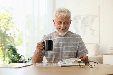 Senior man with cup of drink reading magazine at table indoors