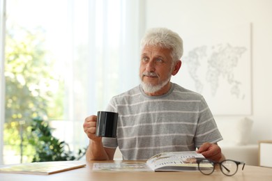 Photo of Senior man with cup of drink reading magazine at table indoors