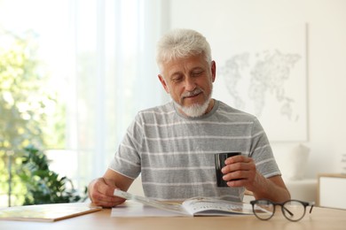 Senior man with cup of drink reading magazine at table indoors