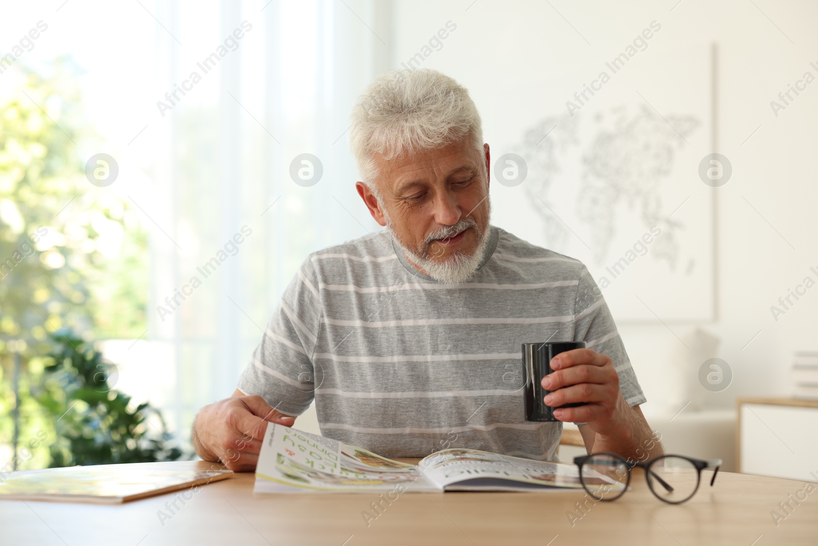 Photo of Senior man with cup of drink reading magazine at table indoors