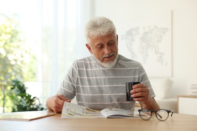 Senior man with cup of drink reading magazine at table indoors