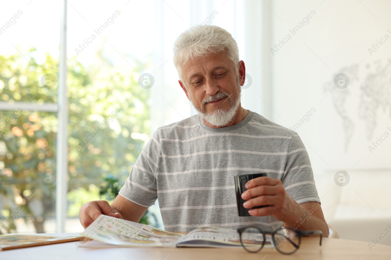 Photo of Senior man with cup of drink reading magazine at table indoors