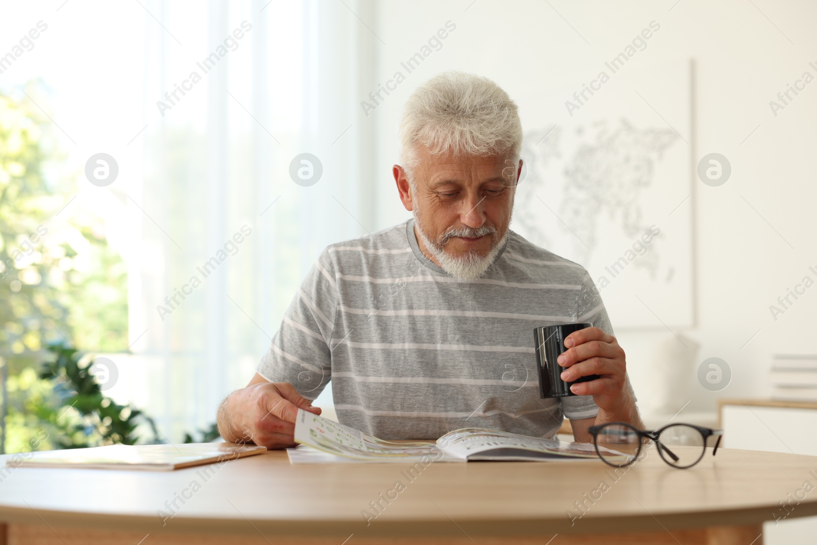 Photo of Senior man with cup of drink reading magazine at table indoors