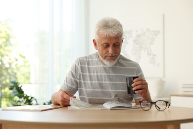 Photo of Senior man with cup of drink reading magazine at table indoors