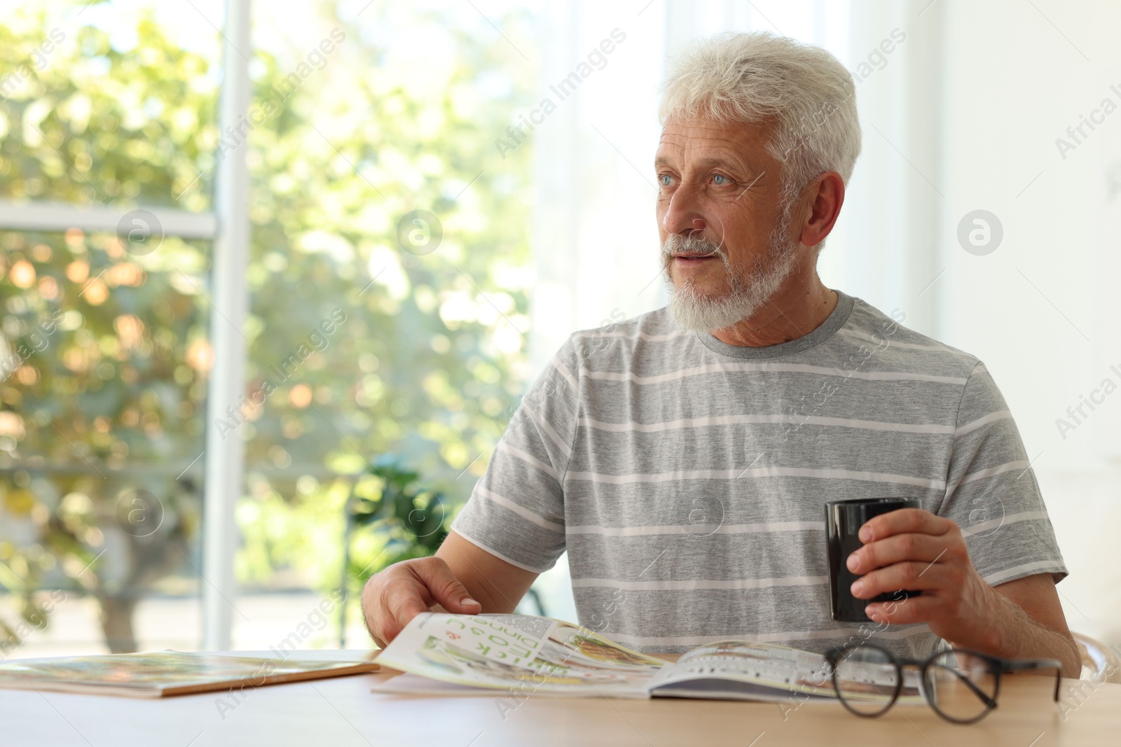 Photo of Senior man with cup of drink reading magazine at table indoors