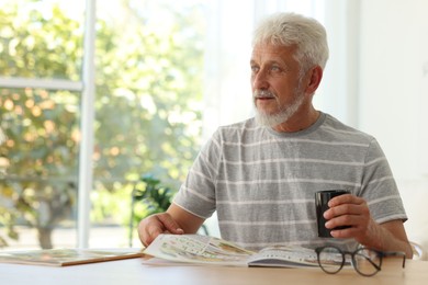 Senior man with cup of drink reading magazine at table indoors