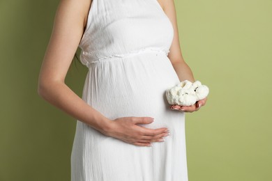 Photo of Young pregnant woman with pair of baby shoes on light olive background, closeup