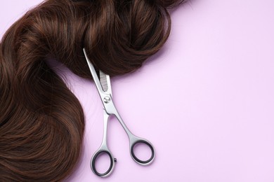 Photo of Brown hair strand and professional scissors on violet background, top view. Space for text