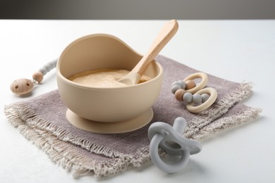 Delicious baby food in bowl with spoon, pacifier and teether on white background