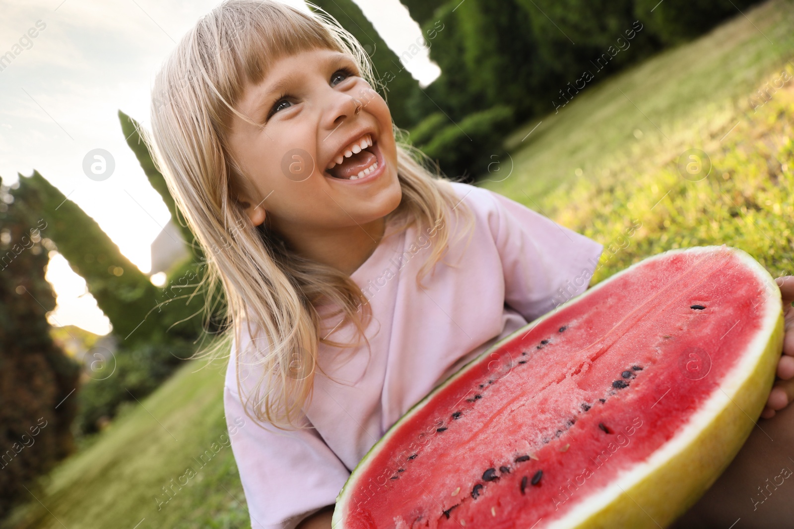 Photo of Cute little girl with half of juicy watermelon on green grass outdoors