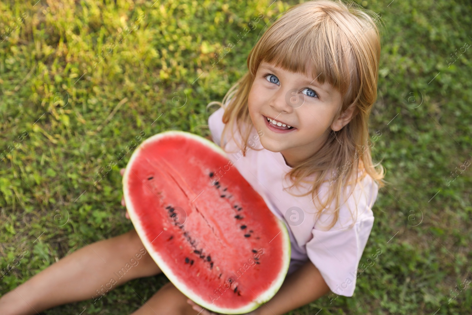 Photo of Cute little girl with half of juicy watermelon on green grass outdoors, above view