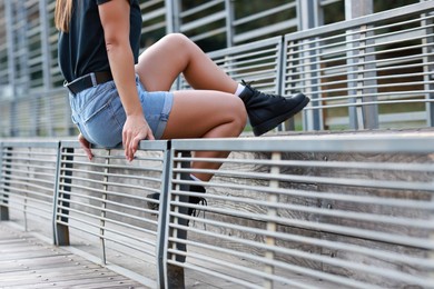 Woman in stylish denim shorts sitting on metal bench backrest outdoors, closeup