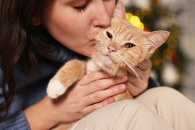 Photo of Woman kissing her cute ginger cat against blurred Christmas lights indoors, closeup