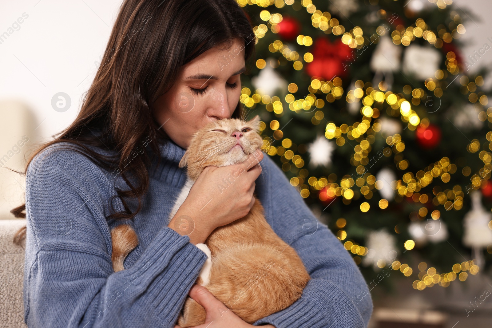 Photo of Woman kissing her cute ginger cat against blurred Christmas lights indoors