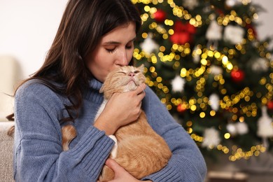 Photo of Woman kissing her cute ginger cat against blurred Christmas lights indoors