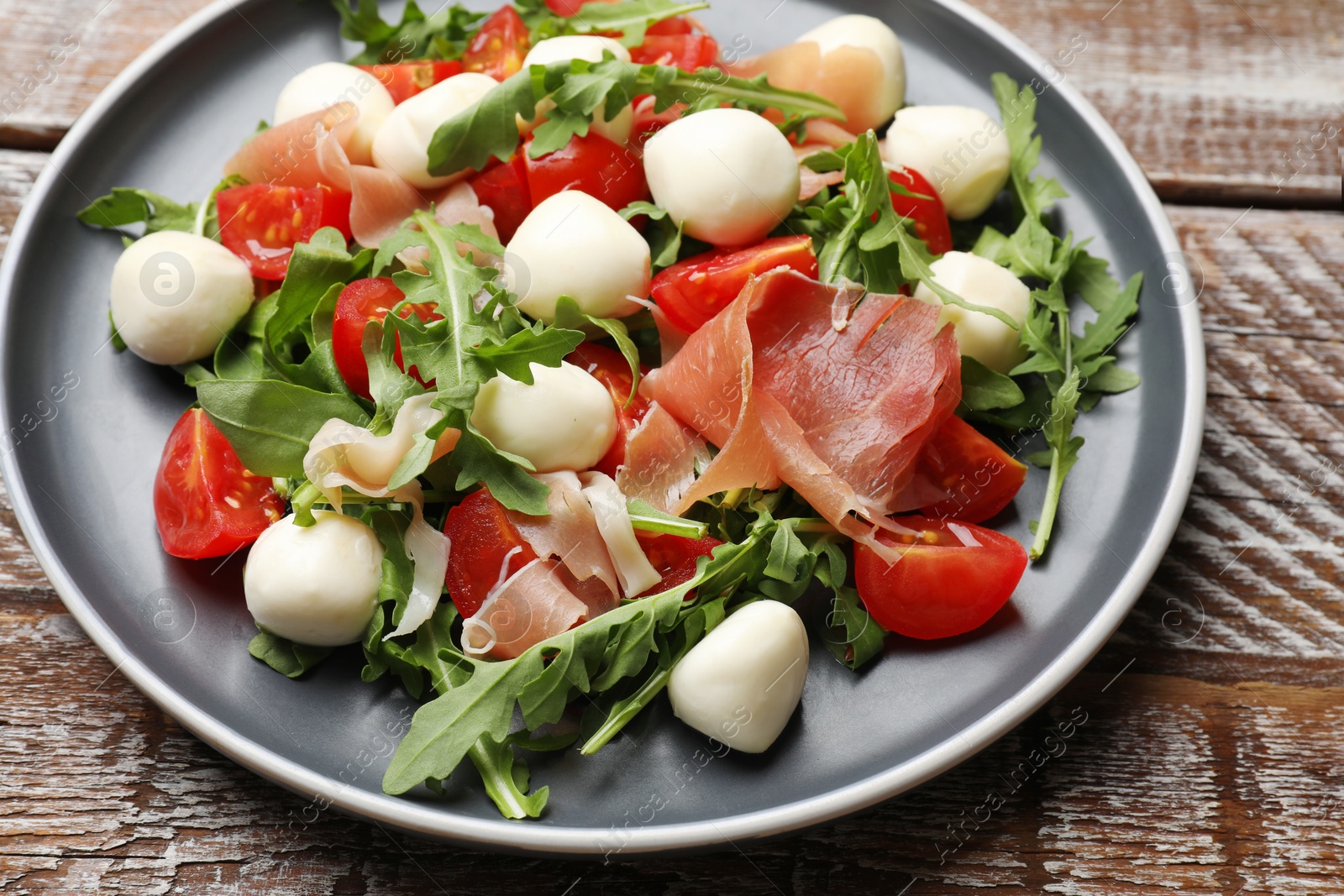 Photo of Tasty salad with arugula, mozzarella cheese, tomatoes and jamon on wooden table, closeup