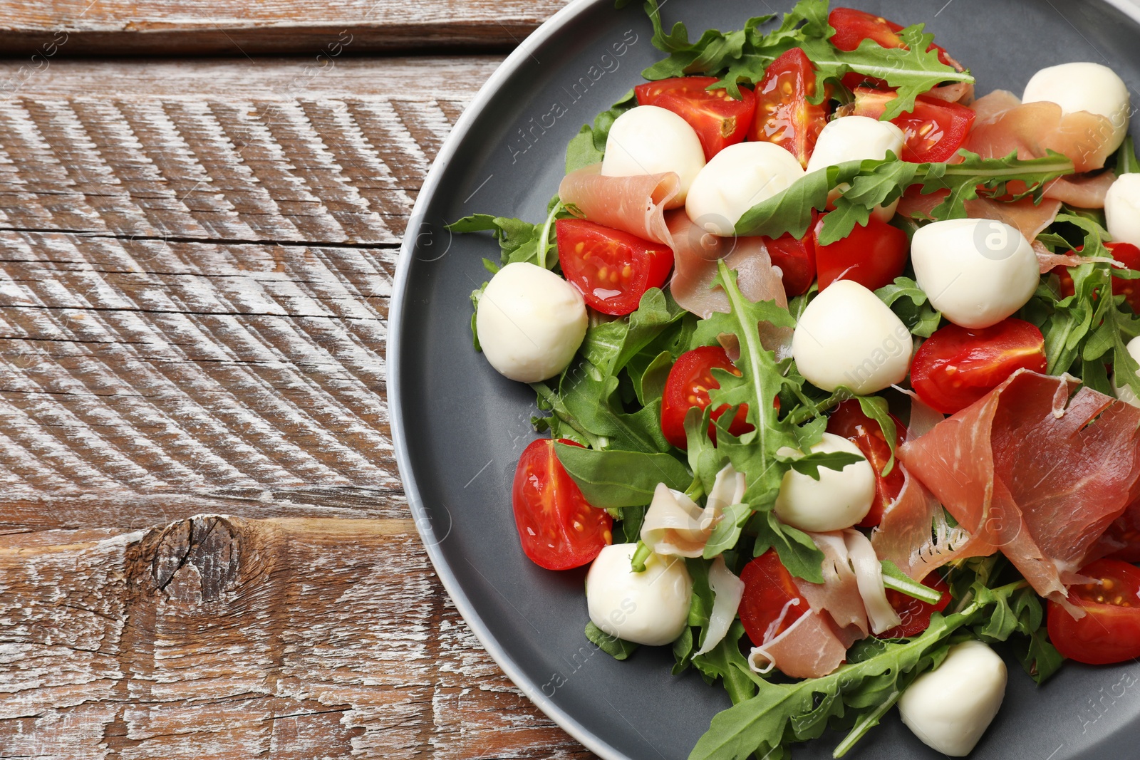 Photo of Tasty salad with arugula, mozzarella cheese, tomatoes and jamon on wooden table, top view. Space for text