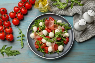 Photo of Tasty salad with arugula, mozzarella cheese, tomatoes and jamon on light blue wooden table, flat lay