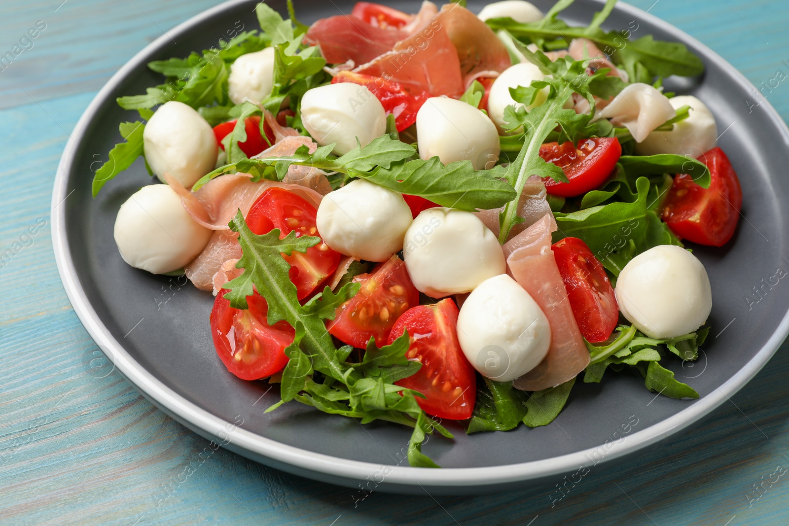 Photo of Tasty salad with arugula, mozzarella cheese, tomatoes and jamon on light blue wooden table, closeup