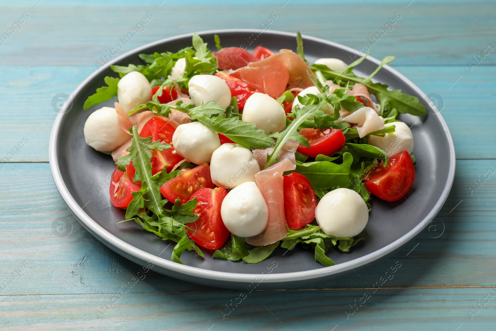 Photo of Tasty salad with arugula, mozzarella cheese, tomatoes and jamon on light blue wooden table, closeup