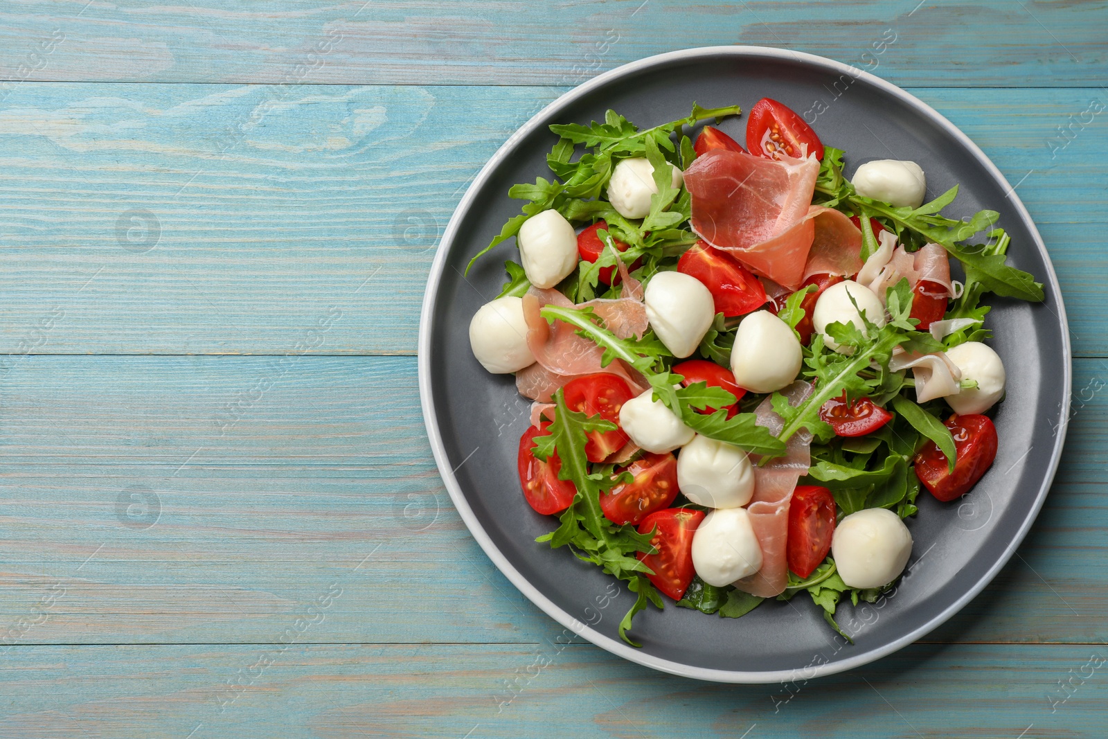 Photo of Tasty salad with arugula, mozzarella cheese, tomatoes and jamon on light blue wooden table, top view. Space for text