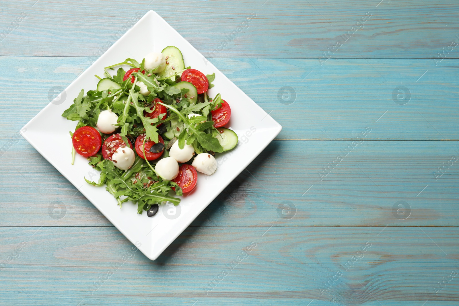 Photo of Tasty salad with arugula, mozzarella cheese and vegetables on light blue wooden table, top view. Space for text