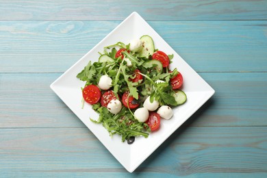 Photo of Tasty salad with arugula, mozzarella cheese and vegetables on light blue wooden table, top view