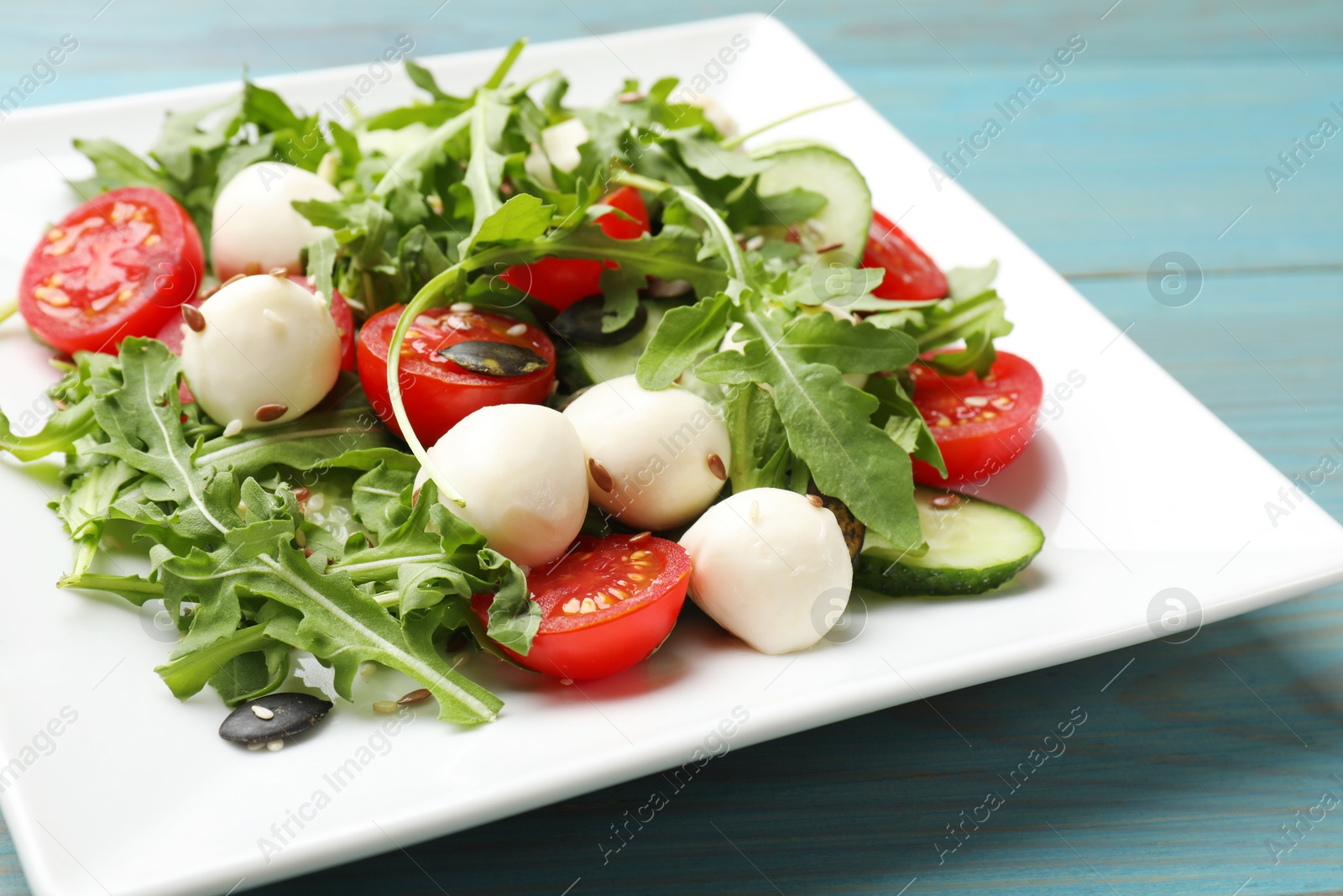 Photo of Tasty salad with arugula, mozzarella cheese and vegetables on light blue wooden table, closeup