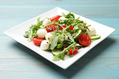 Photo of Tasty salad with arugula, mozzarella cheese and vegetables on light blue wooden table, closeup