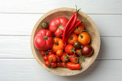 Different ripe tomatoes and chili peppers in bowl on white wooden table, top view