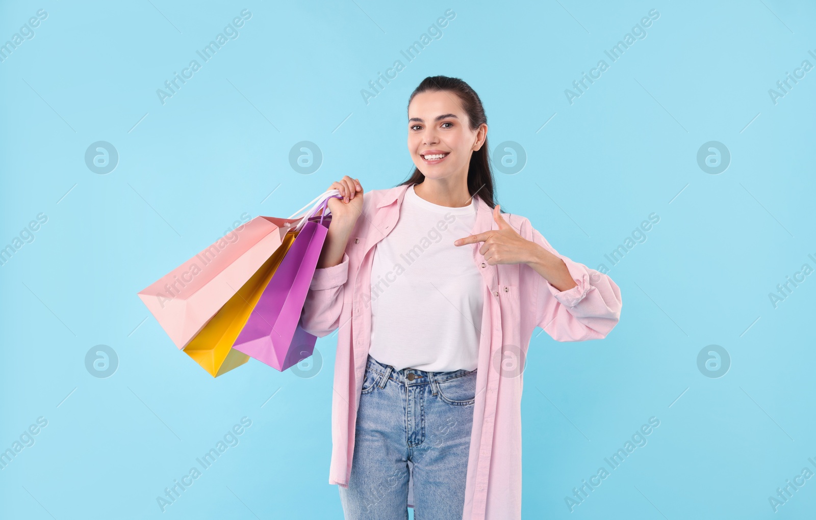 Photo of Smiling woman pointing at colorful shopping bags on light blue background