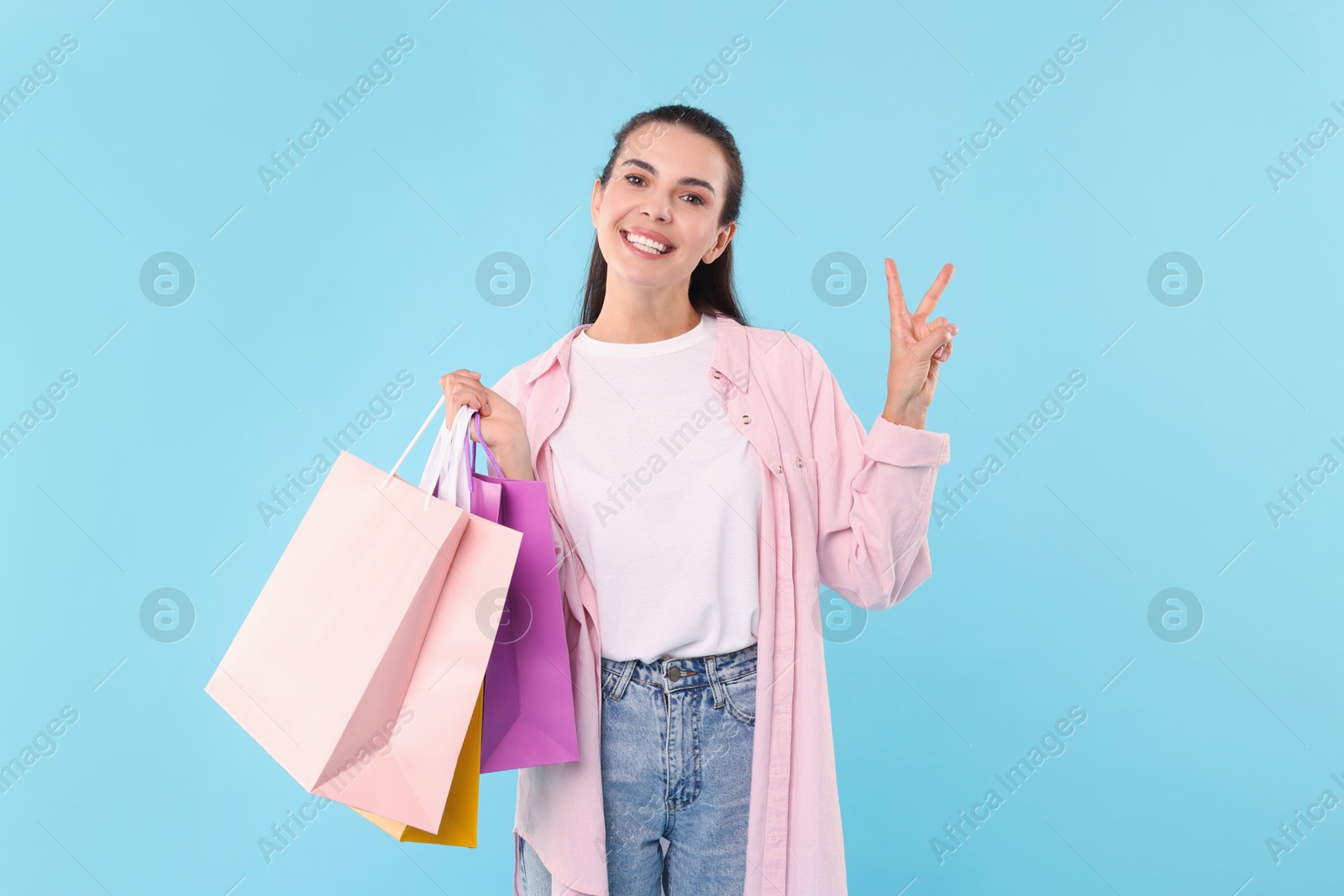 Photo of Smiling woman with colorful shopping bags showing peace sign on light blue background