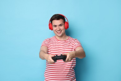 Happy young man in headphones playing video games with controller on light blue background