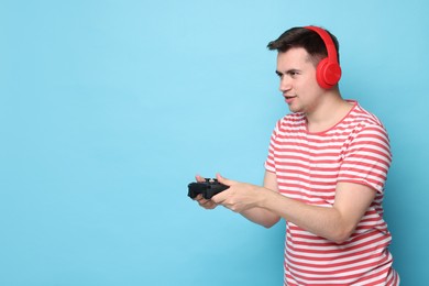 Young man in headphones playing video games with controller on light blue background, space for text