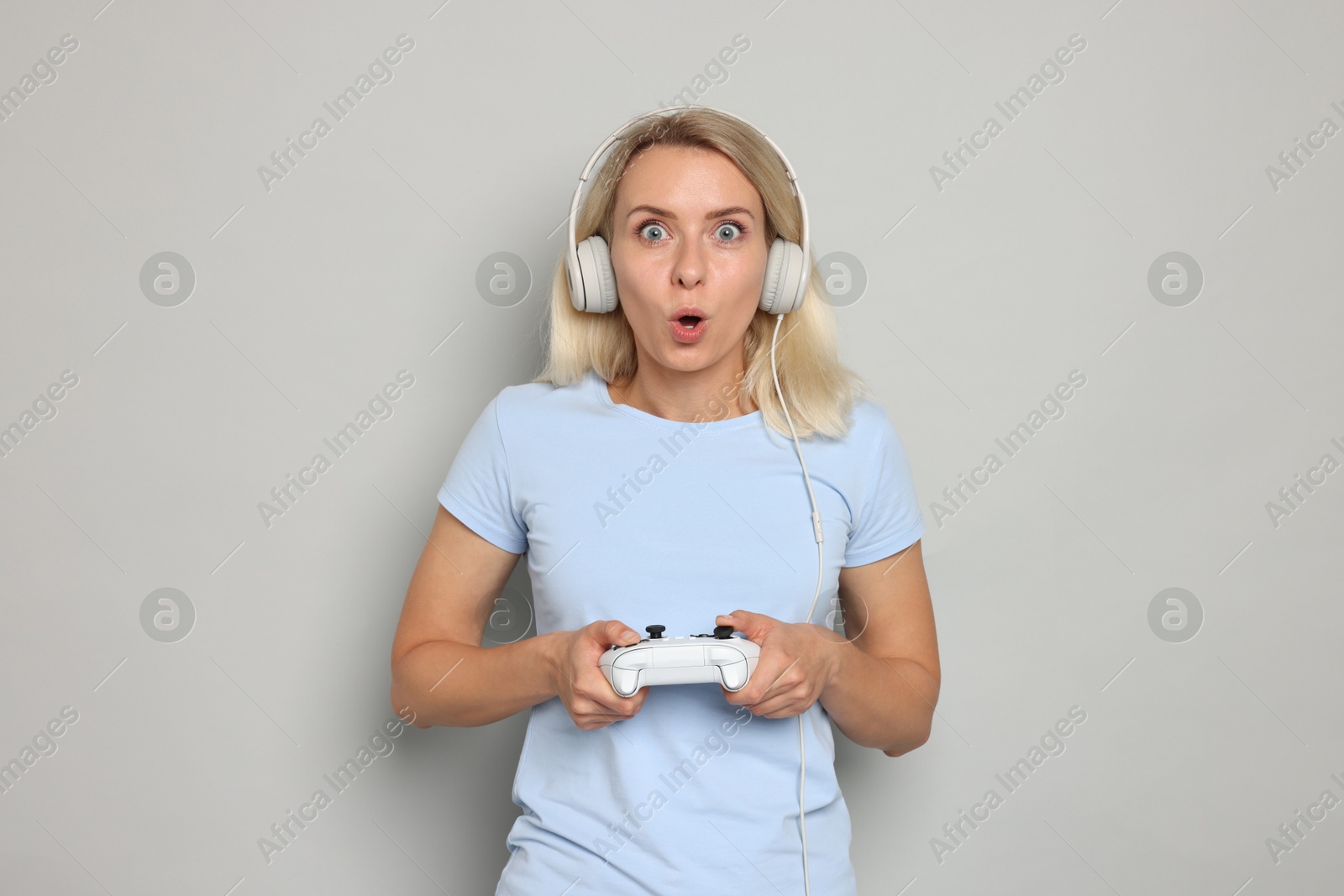 Photo of Surprised woman in headphones playing video games with controller on gray background