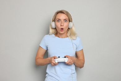 Surprised woman in headphones playing video games with controller on gray background