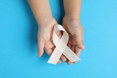 Photo of Child with white ribbon on light blue background, top view. Bone cancer and lung disease awareness