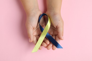 Photo of Child with yellow and blue ribbon on pink background, top view. Down syndrome awareness