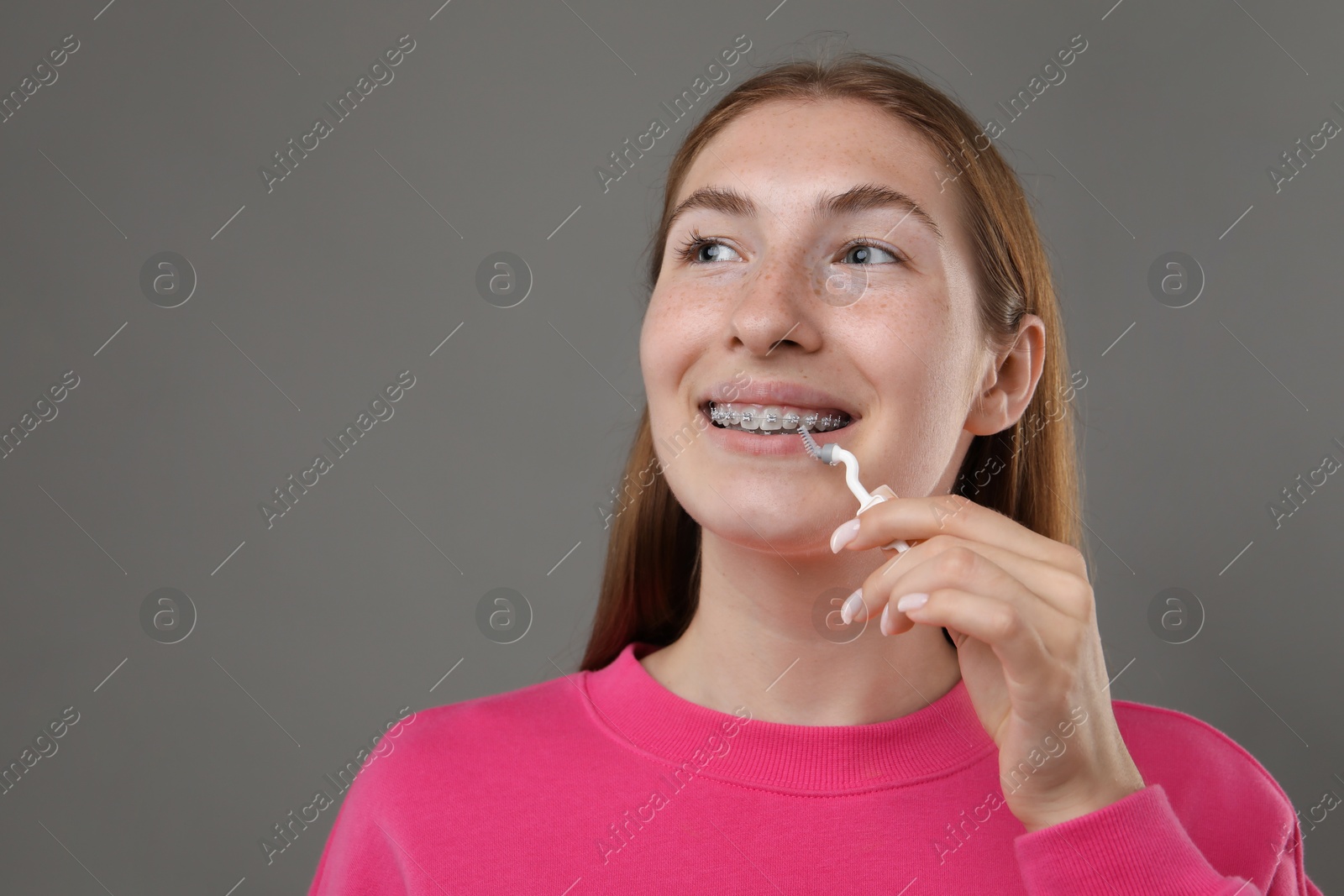 Photo of Smiling girl with braces cleaning teeth with interdental brush on grey background