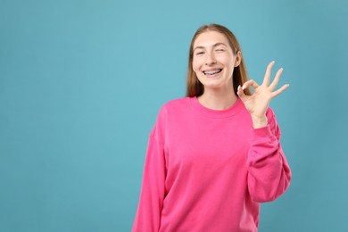 Photo of Smiling girl with braces showing ok gesture on light blue background, space for text