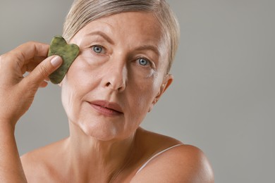 Beautiful woman doing facial massage with gua sha tool on grey background, closeup