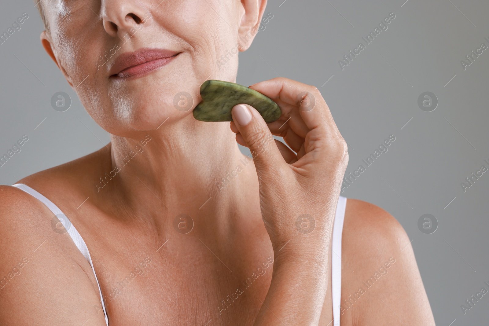 Photo of Woman doing facial massage with gua sha tool on grey background, closeup