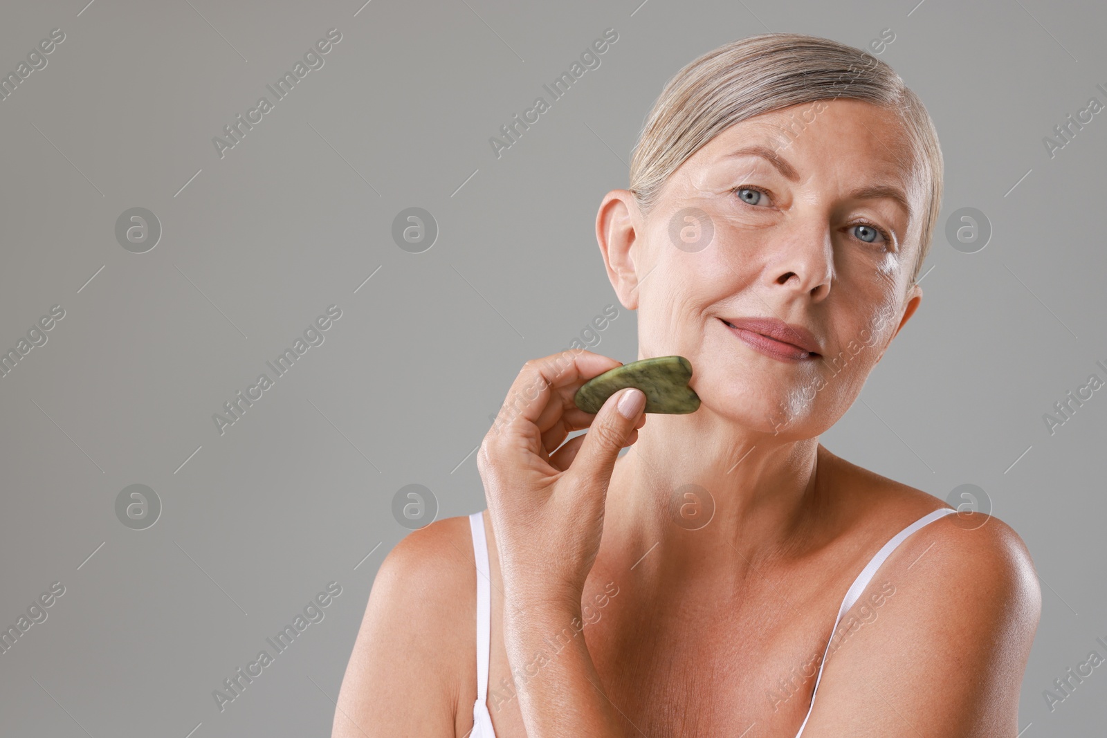 Photo of Beautiful woman doing facial massage with gua sha tool on grey background, space for text