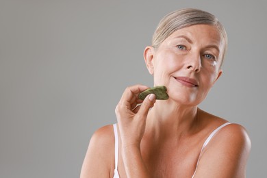 Beautiful woman doing facial massage with gua sha tool on grey background, space for text