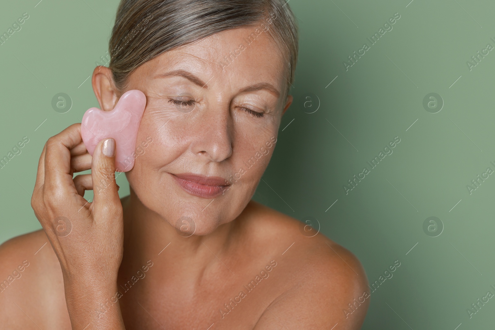 Photo of Beautiful young woman doing facial massage with gua sha tool on light green background