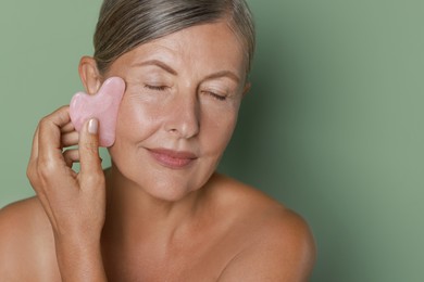 Beautiful young woman doing facial massage with gua sha tool on light green background