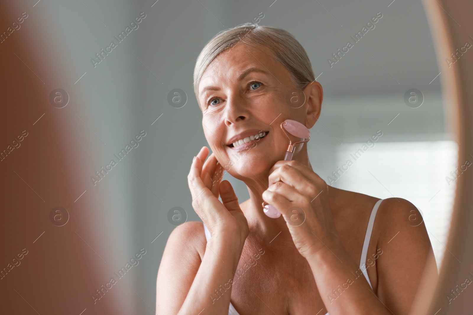 Photo of Beautiful woman doing facial massage with roller near mirror at home