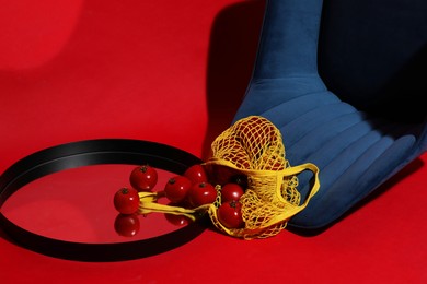 Photo of Creative still life with overturned dark blue armchair, net bag, tomatoes and mirror on red background