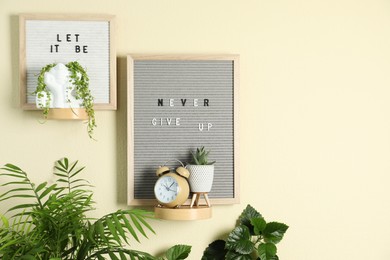 Photo of Letter boards with phrases Let It Be, Never Give Up, alarm clock and plants on beige wall. Space for text