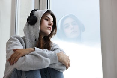 Loneliness concept. Sad teenage girl in headphones listening to music near window at home, low angle view